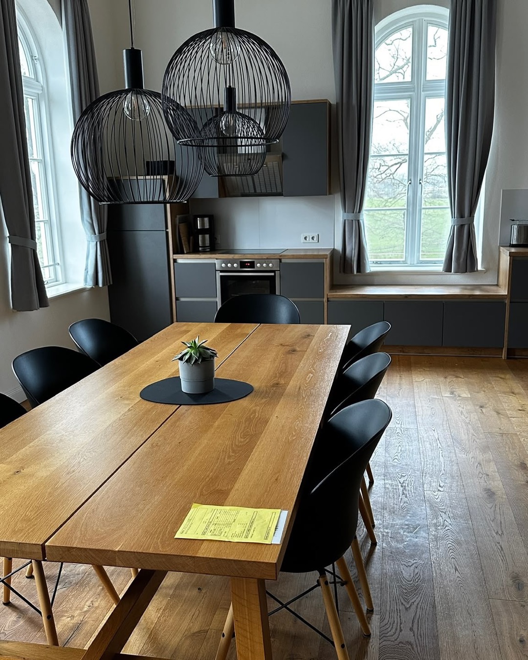 Der Speiseraum mit Küche, langem Holztisch und hohen Bogenfenstern