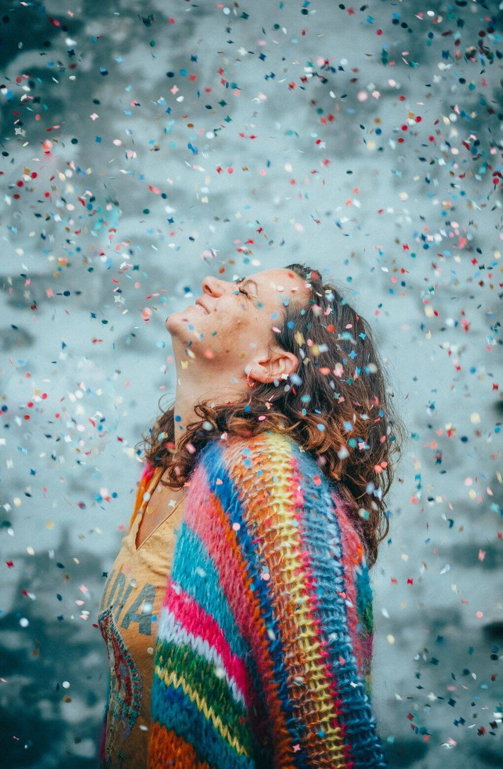 Julia hebt lachend das Gesicht in einen Regen aus buntem Konfetti, um die Schultern ein bunter Strickcardigan