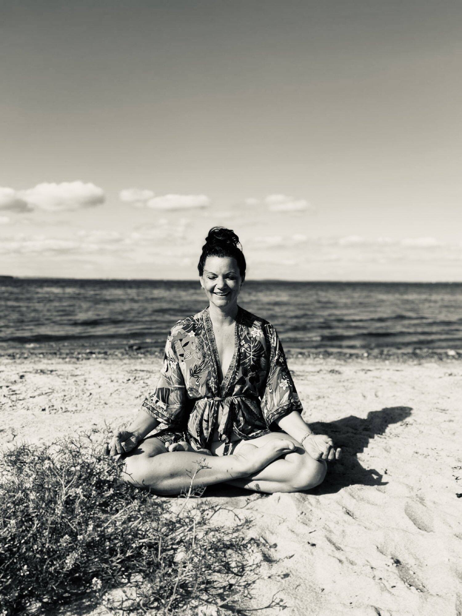 Julia meditiert im Schneidersitz am Strand, hinter ihr das offene Meer