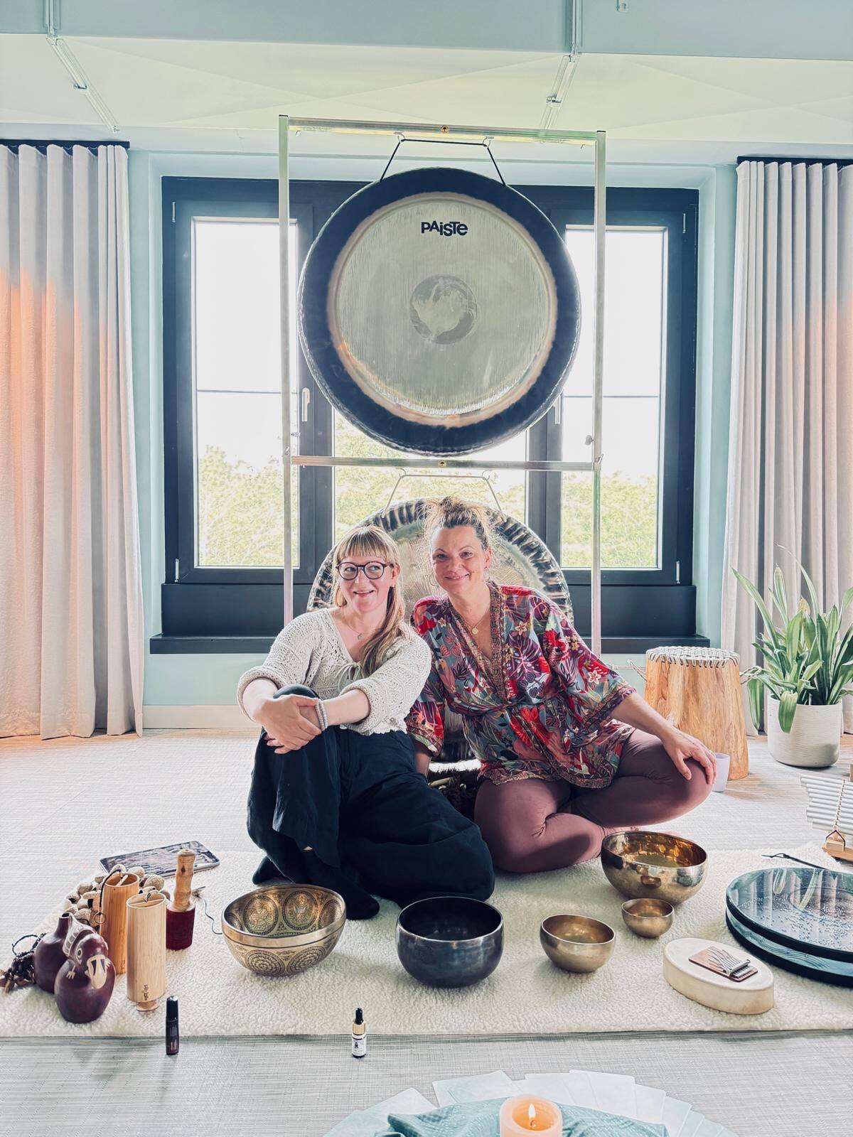 Jurga und Julia sitzen lächelnd vor einem großen Gong, vor ihnen Klangschalen