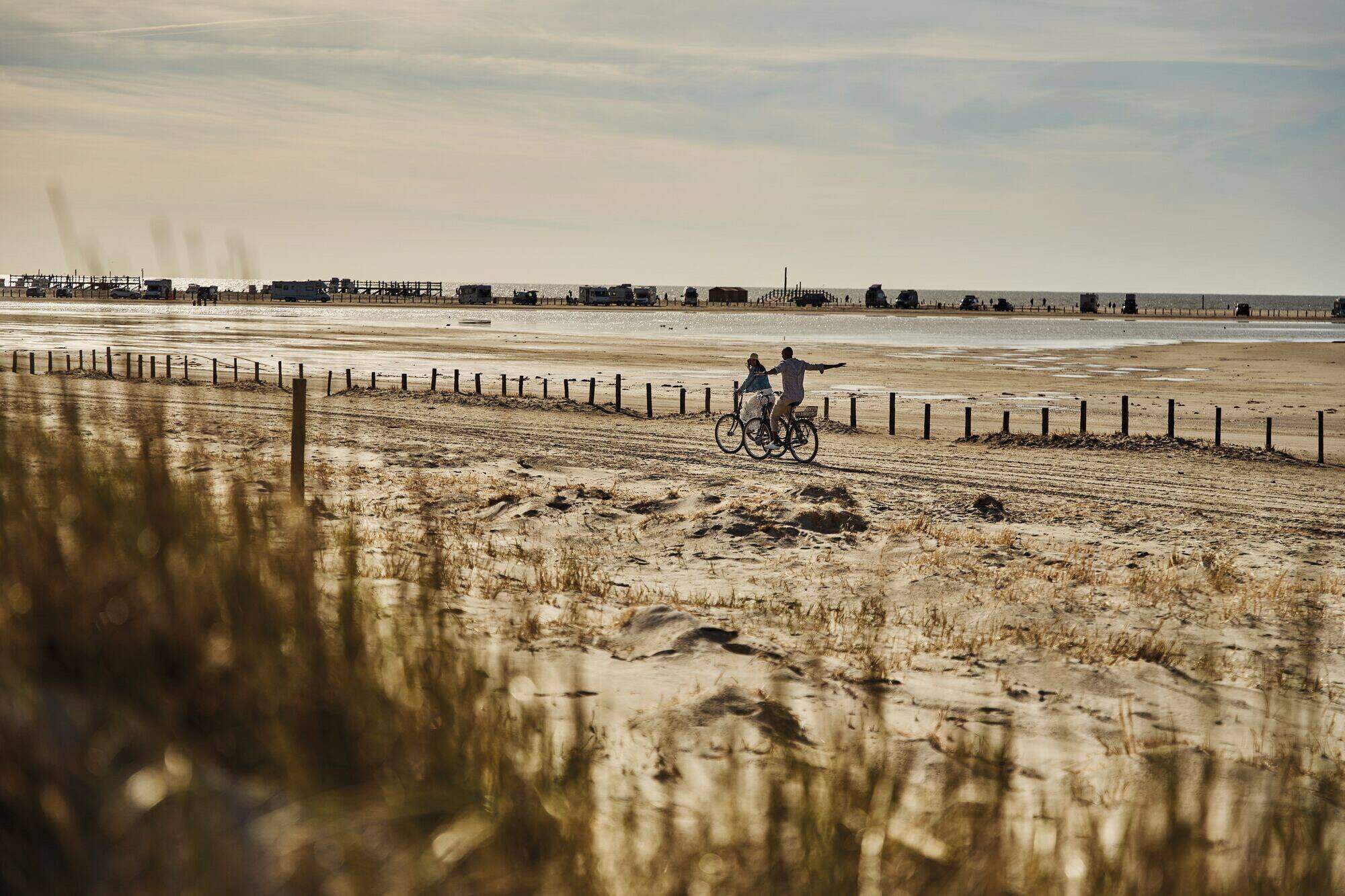 Weiter Strand von Sankt Peter-Ording im warmen Licht, zwei Radfahrende mit ausgebreiteten Armen
